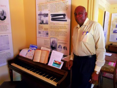 Alfred Rankins stands next to a historical piano in Latimer House which can now be played by visitors.