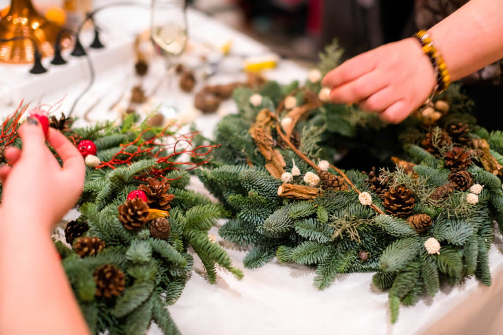 Christmas wreath weaving workshop. Woman hands decorating holiday wreath made of spruce branches, cones and various organic decorations on the table