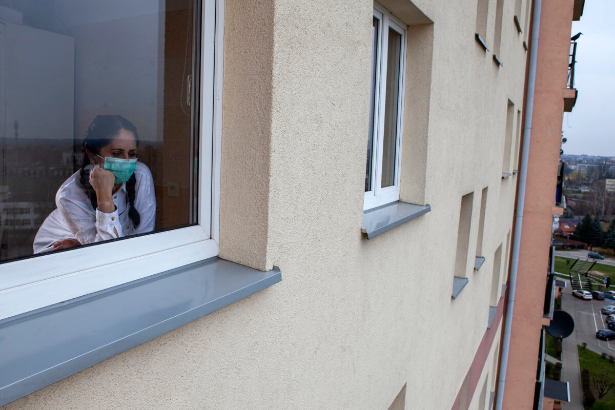A 40-year-old woman wearing a protective mask is looking out of the window. Home quarantine for 14 days due to the coronavirus COVID-19 epidemic.