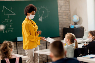 Happy teacher giving exam paper to her student while wearing protective face mask in the classroom.