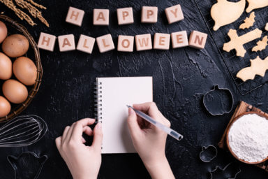 Young woman is reading and writing recipe of making Halloween cookies, design concept of preparing for Halloween party, top view, flat lay, overhead.