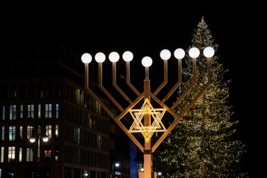 Huge illuminated menorah with round lamps and lighted Jewish symbol David’s star on blurry background of Christmas tree in Berlin Germany. Night cityscape with Hanukah decorations