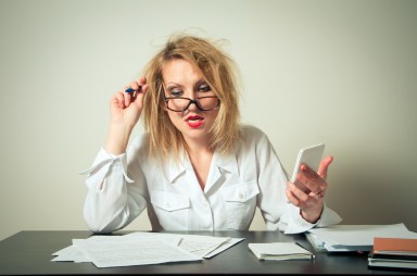 Irritated Businesswoman With Tousled Hair Analyzing Documents At Desk Against Gray Background