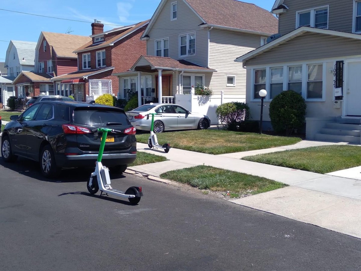 Two green e-scooters block a residential sidewalk and the back of a black vehicle.