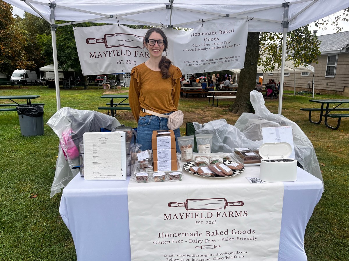 Woman in a mustard colored shirt stands in front of booth that says Mayfield Farms at a festival