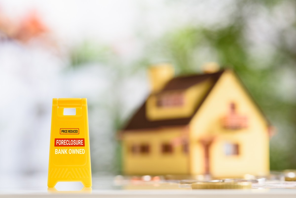 Foreclosures and foreclosed home for sale property listings, financial concept : Yellow warning sign board with the words FORECLOSURE FOR SALE BANK-OWNED, a two-story model house, coins on a table.