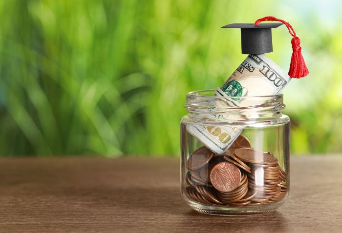 Scholarship concept. Glass jar with coins, dollar banknotes and graduation cap on wooden table, space for text